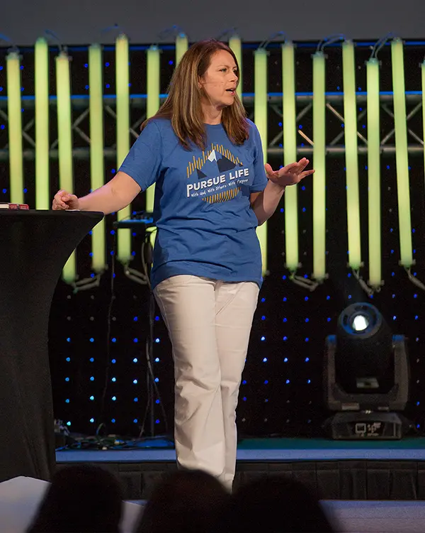 Lisa Anderson presenting on stage wearing a “Pursue Life” event t-shirt and white pants with a modern lighted backdrop.